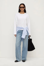 Woman wearing a white blouse, light blue jeans, and black sunglasses against a white background