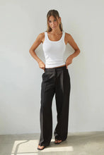 Woman wearing a white tank top and black pants against a plain background