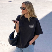 Woman wearing a black t-shirt with a logo, holding a phone, and carrying a blue bag on a light-colored pavement.