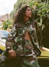 Person wearing a camouflage outfit standing next to a yellow car with greenery in the background