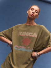 Person wearing a green oversized t-shirt with 'KINGA CSILLA' print against a blue background